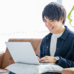 明るいリビングのソファで、クッションの上に置いたノートパソコンを笑顔で操作する若い男性。