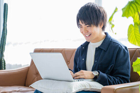 明るいリビングのソファで、クッションの上に置いたノートパソコンを笑顔で操作する若い男性。