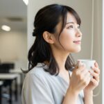 明るいオフィスの窓際で、女性が少し考え事をしながらも、希望に満ちた表情でマグカップを持って立っている