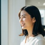 明るい自然光が差し込むオフィスで、女性が窓の外を見つめ、穏やかな表情で微笑んでいる。