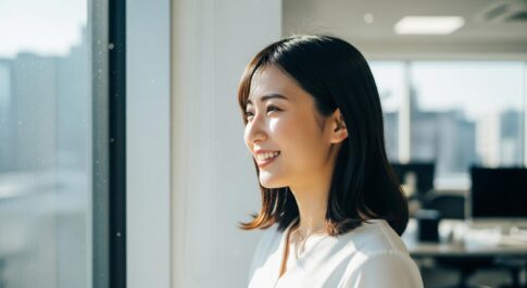 明るい自然光が差し込むオフィスで、女性が窓の外を見つめ、穏やかな表情で微笑んでいる。