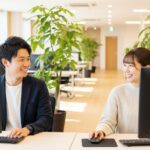 明るく清潔感のあるオフィスで、男性と女性が笑顔でパソコン作業をしながら穏やかに会話している。