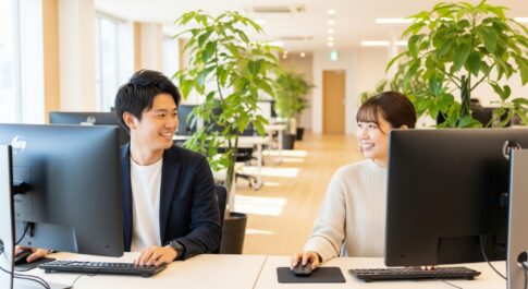 明るく清潔感のあるオフィスで、男性と女性が笑顔でパソコン作業をしながら穏やかに会話している。