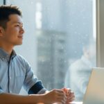 明るいオフィスカジュアルの服装をした男性が、ノートパソコンの前で、満足げな表情で窓の外を見ている