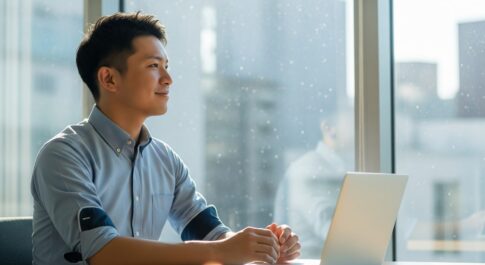 明るいオフィスカジュアルの服装をした男性が、ノートパソコンの前で、満足げな表情で窓の外を見ている